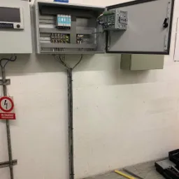 Otwarta szara szafa sterownicza z widocznymi elementami elektrycznymi, zamontowana na ścianie, z pionowo poprowadzonymi kablami w korytku, obok tabliczka ostrzegawcza o napięciu.