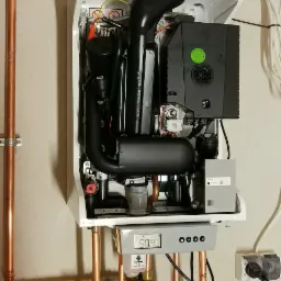 Zamontowany, biały piec gazowy z widocznymi rurami miedzianymi i zaworami, wyświetlacz pokazuje 50 stopni Celsjusza, podłączony do gniazdka elektrycznego.