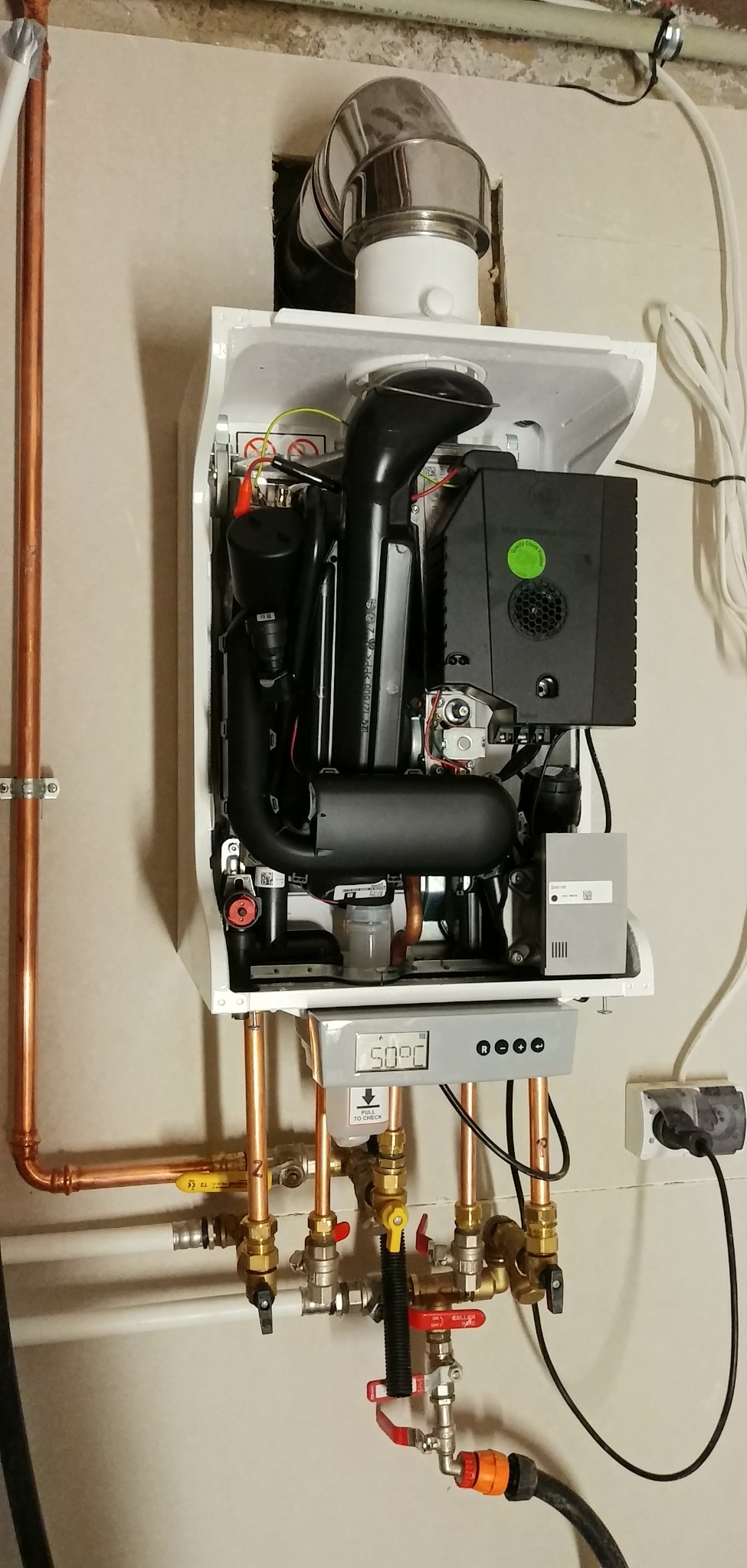 Zamontowany, biały piec gazowy z widocznymi rurami miedzianymi i zaworami, wyświetlacz pokazuje 50 stopni Celsjusza, podłączony do gniazdka elektrycznego.