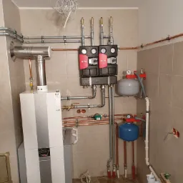 Nowoczesny, kondensacyjny piec gazowy Wolf z szarymi pasami, zainstalowany w pomieszczeniu z beżowymi płytkami, widoczne rury miedziane i stalowe, manometry, oraz zbiorniki wyrównawcze w kolorze...