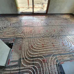 Białe rury ogrzewania podłogowego ułożone spiralnie na czerwonej siatce w surowym wnętrzu, widok z góry na instalację przed zalaniem wylewką, w tle okno z widokiem na plac budowy.