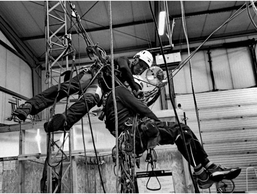 Czarno-białe zdjęcie dwóch osób w uprzężach, podczas szkolenia z technik dostępu linowego w hali z metalową konstrukcją i wentylatorem w tle.
