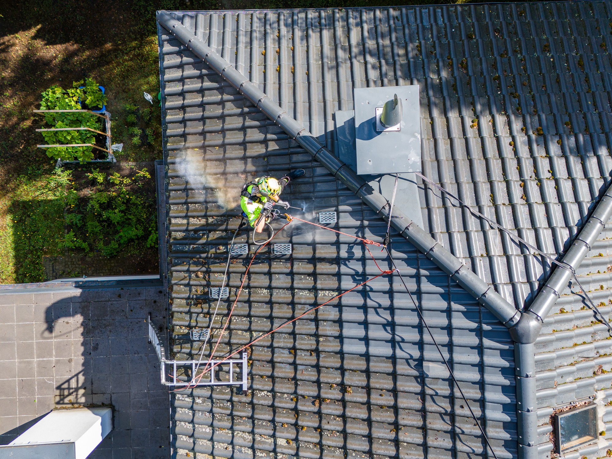 Pracownik w uprzęży, z góry, czyści dach z szarej dachówki za pomocą myjki ciśnieniowej. Widoczne zacieki i mech. Obok zieleń i drabina na linie.