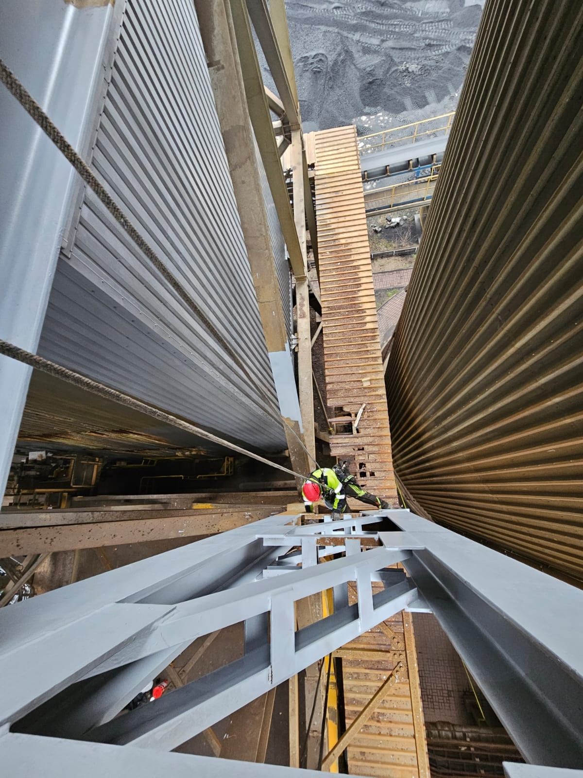 Pracownik w uprzęży, na wysokości, maluje metalową konstrukcję przemysłową na szaro. Widok z góry, perspektywa podkreśla skalę i trudność zadania.