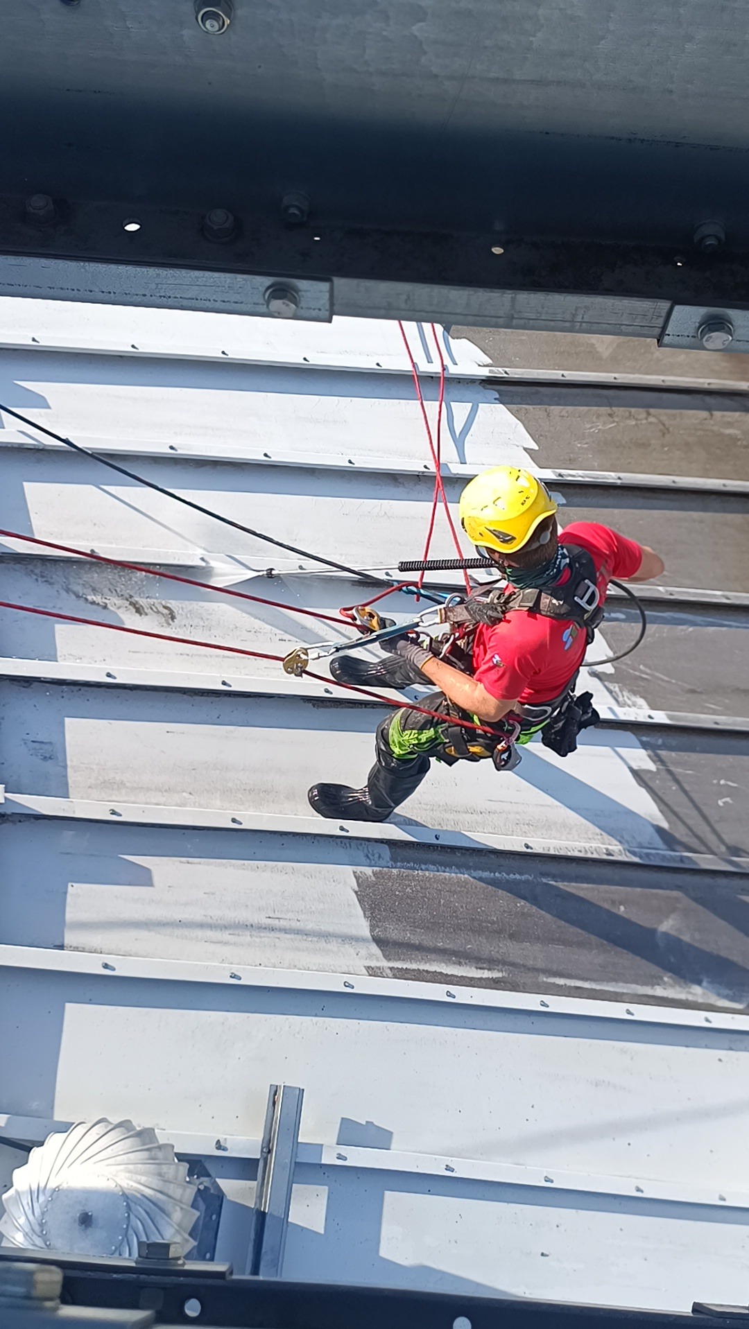 Pracownik w kasku, zabezpieczony linami, wykonuje prace na wysokości na metalowej konstrukcji. Widoczne ślady mycia, co sugeruje czyszczenie ciśnieniowe.