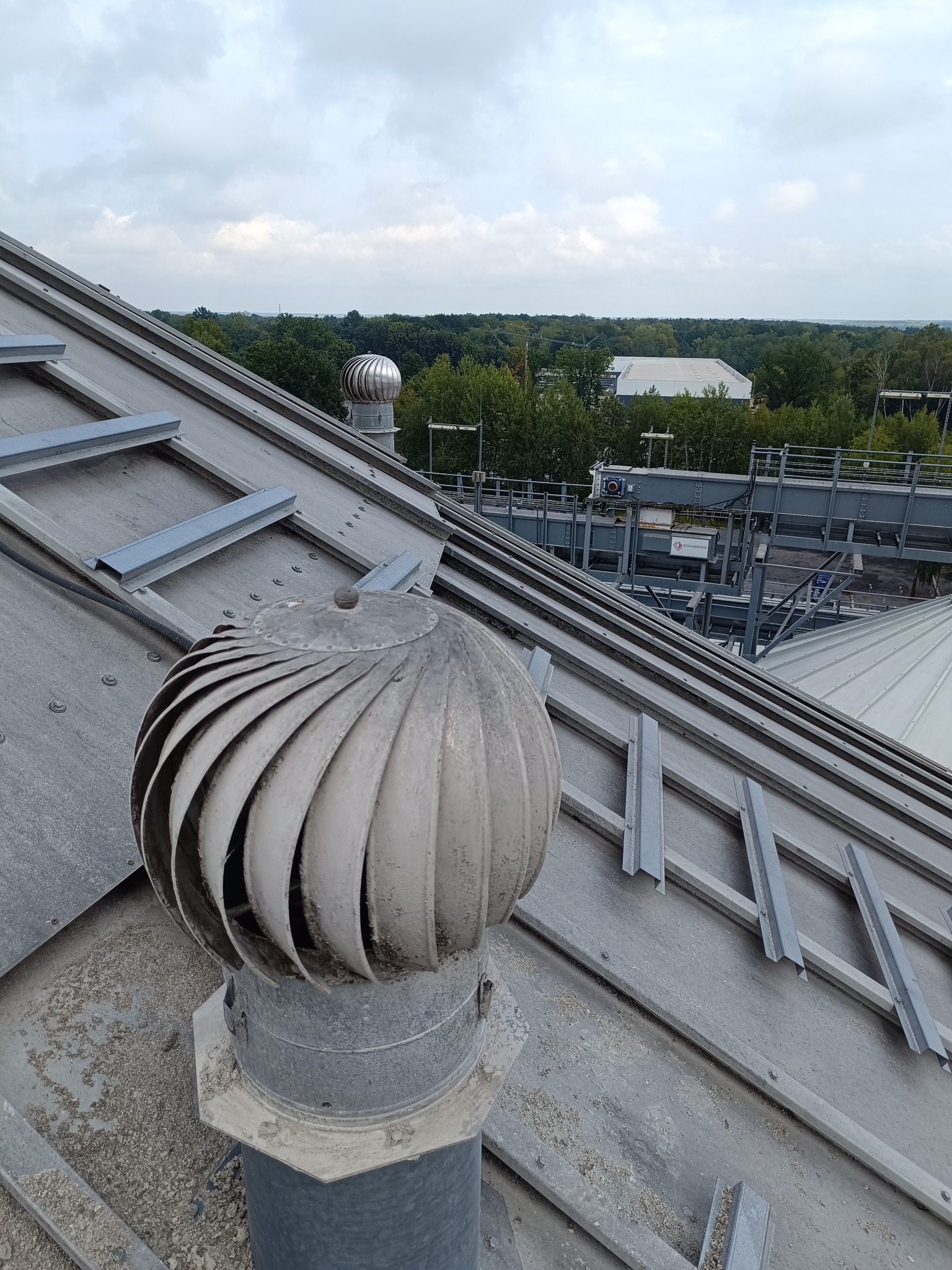 Z bliska: zabrudzony, metalowy wentylator turbinowy na dachu zbiornika. Widoczne zanieczyszczenia i elementy konstrukcyjne dachu. W tle zieleń i budynki.