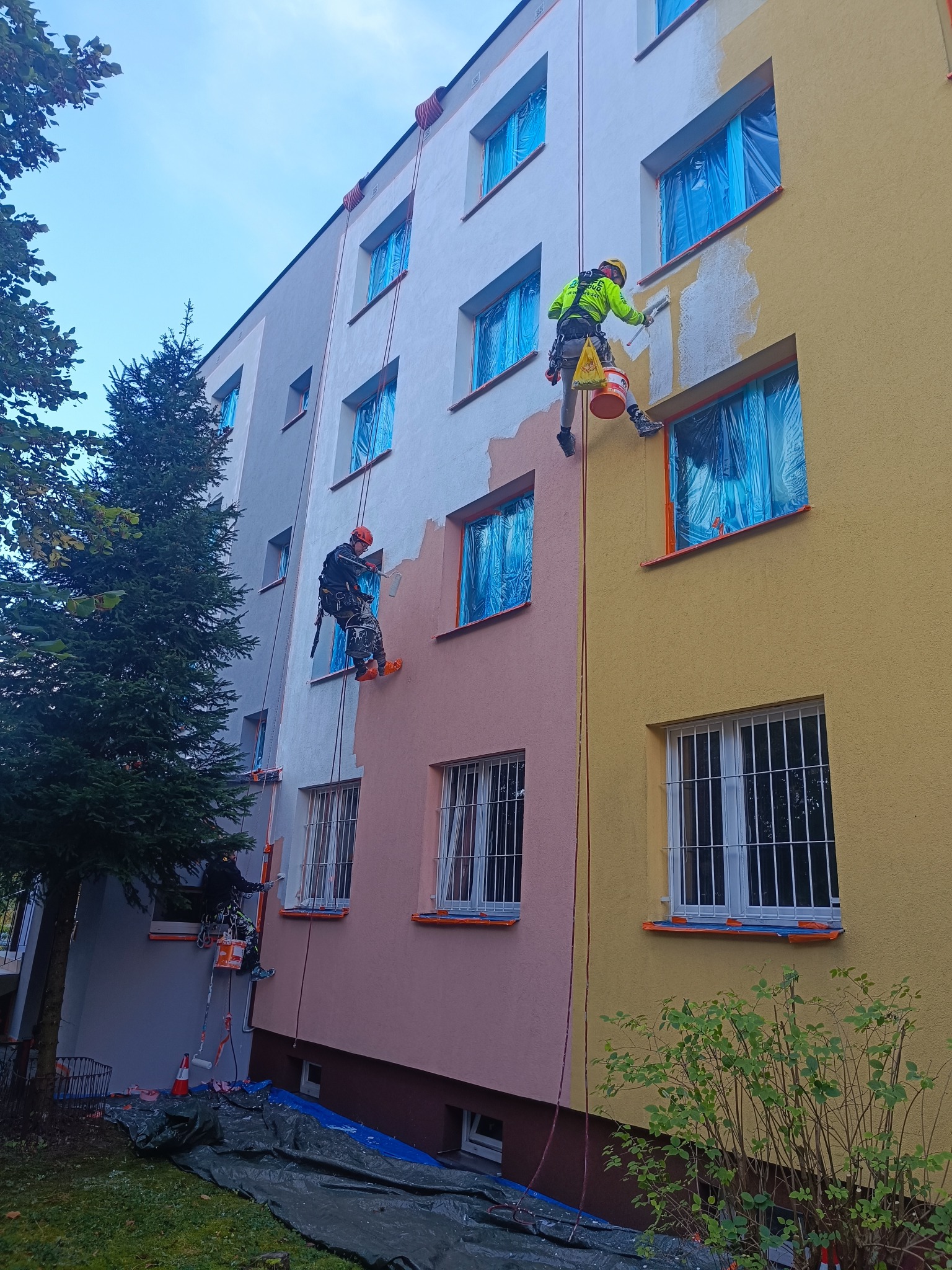 Dwóch alpinistów w trakcie nakładania farby na elewację bloku, okna zabezpieczone folią. Widoczne fragmenty pomalowanej i niepomalowanej ściany w różnych kolorach.