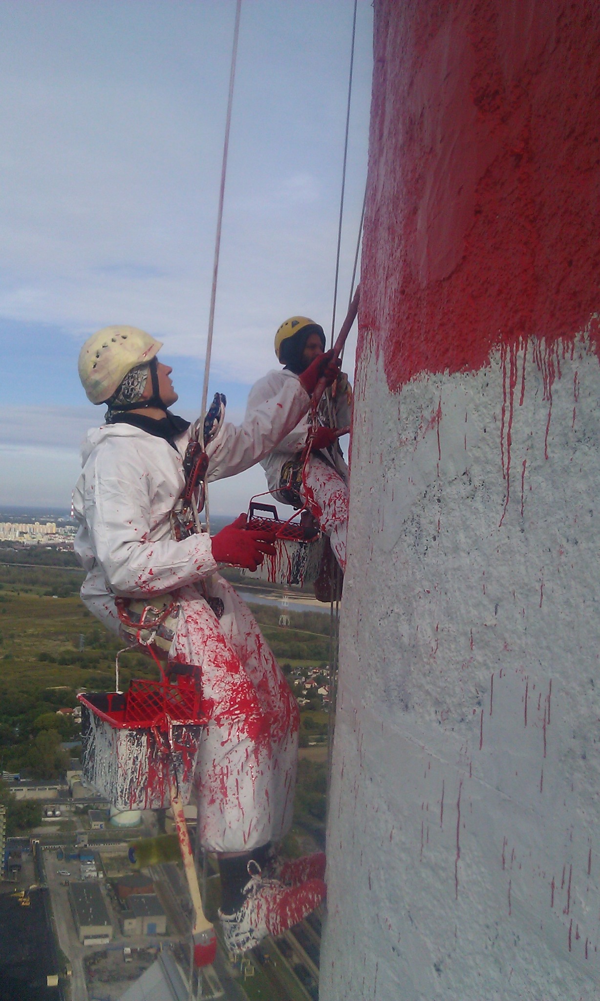 Dwóch alpinistów w kombinezonach maluje na czerwono wysoki komin przemysłowy, zabezpieczeni linami. Widok z góry na miasto w tle.