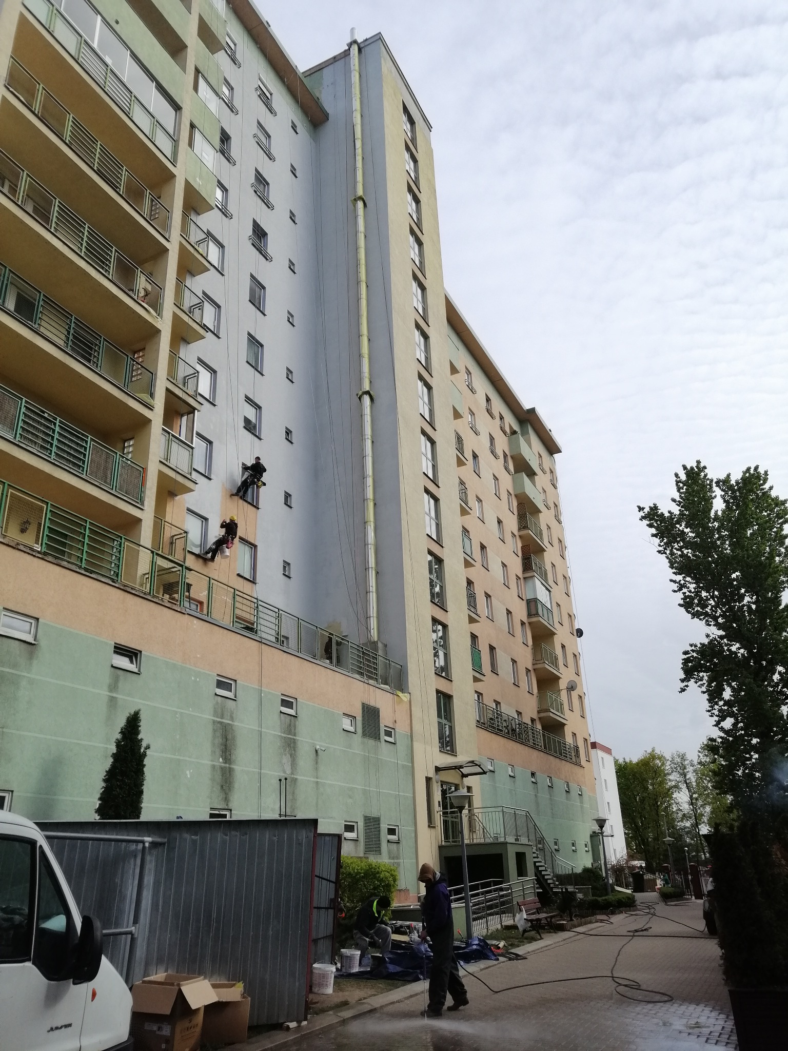 Dwóch alpinistów na linach czyści elewację bloku. Na dole pracownik z myjką ciśnieniową. Widoczne ślady zabrudzeń na elewacji. Ujęcie z dołu, pochmurne niebo.