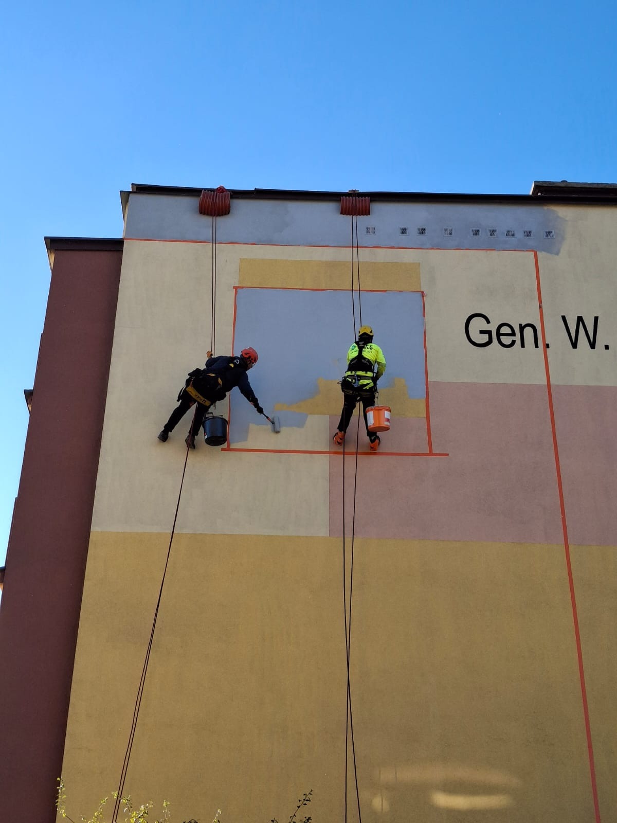 Dwóch alpinistów w uprzężach maluje fragment elewacji budynku na wysokości, używając wałków. Widoczne liny, farba i kolorowe bloki na ścianie. Praca w toku.