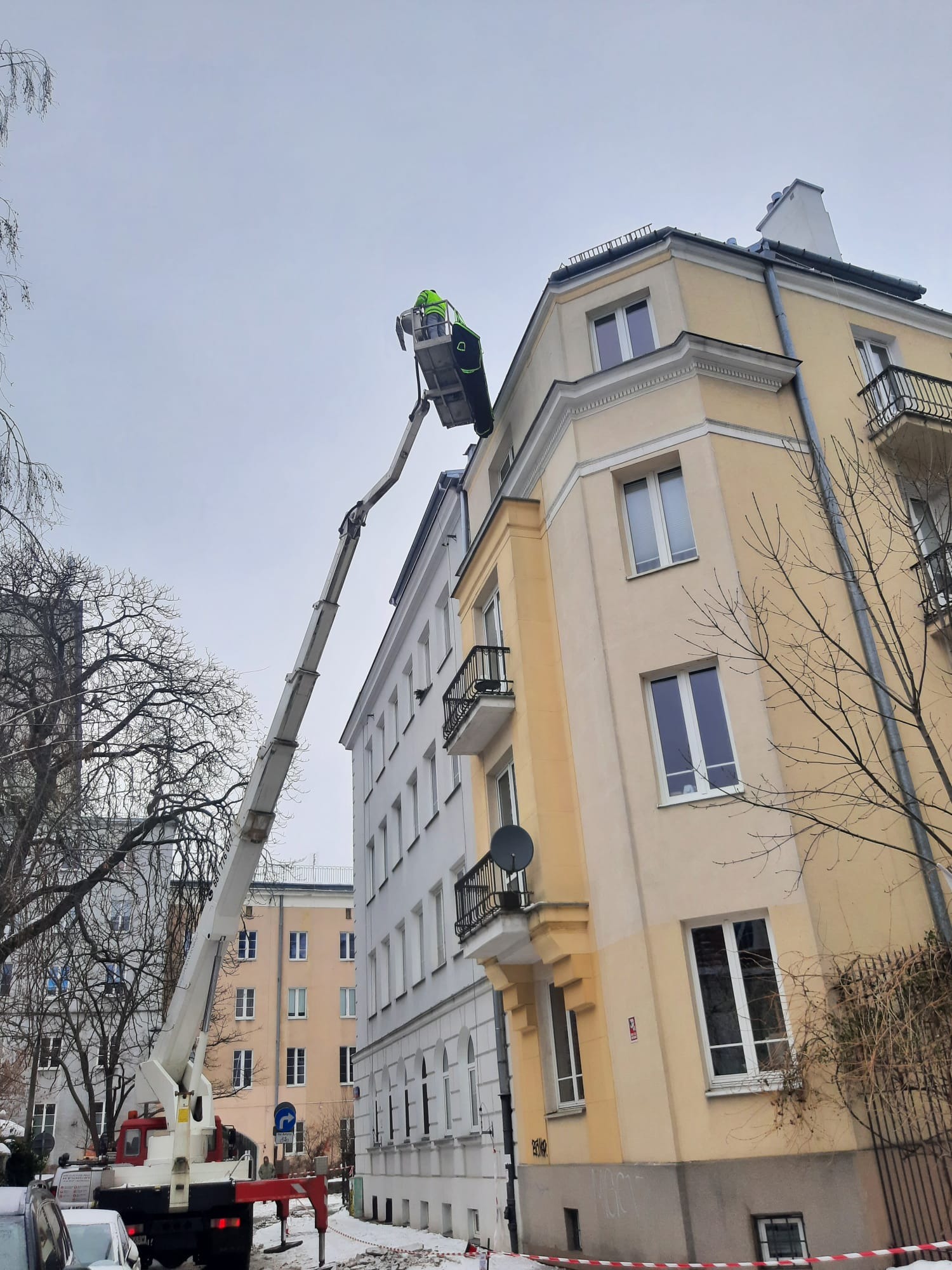 Pracownik w koszu podnośnika teleskopowego przy elewacji kamienicy w zimowej scenerii. Widok z dołu na żółtą i białą fasadę budynku.