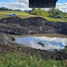 Wykopany staw z wodą i odbiciem nieba, na brzegu ziemia i roślinność, w tle biała ciężarówka i budynek. Nad wykopem fragment łyżki koparki.