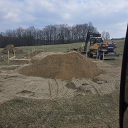 Usługi transportowe Konkret - Żółta koparka Volvo na terenie przygotowywanym pod budowę, z dużą hałdą urobku, wyraźnymi śladami opon i drewnianymi palikami wyznaczającymi obszar prac, pod pochmurnym niebem.
