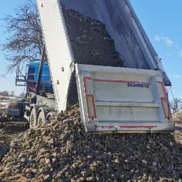 Ciężarówka z podniesioną naczepą wysypuje pokruszony materiał budowlany na ziemię, widoczne ślady opon w błocie, błękitne niebo w tle.