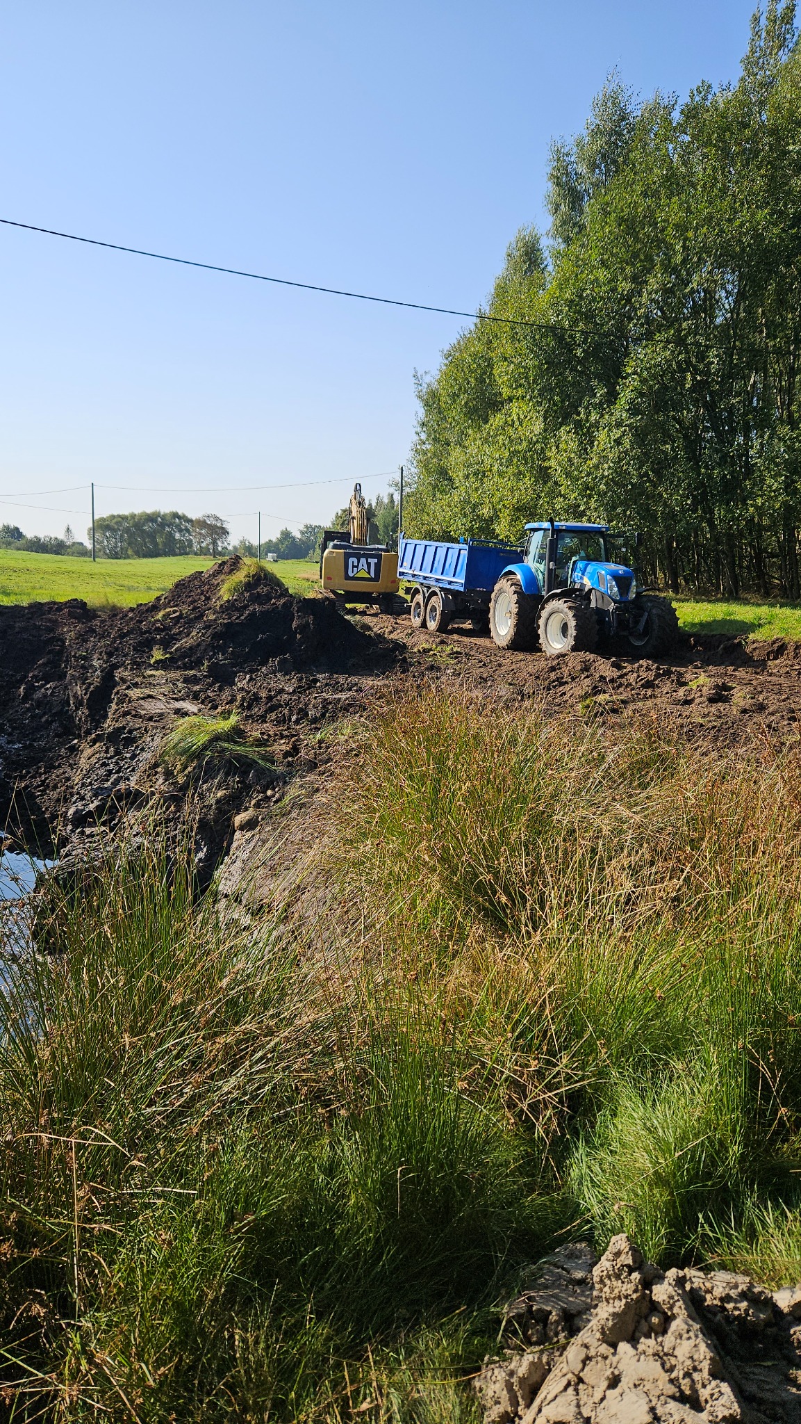 Błękitny traktor z przyczepą i koparka CAT na skraju wykopu z wodą, w tle zieleń drzew i łąk, słoneczny dzień, widok z poziomu gruntu.