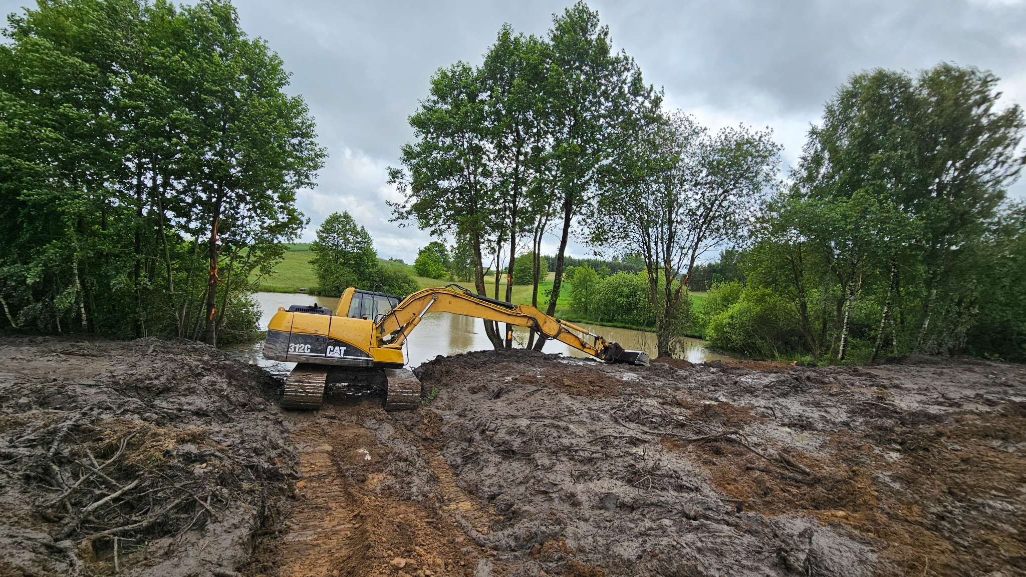 Żółta koparka CAT 312C pracuje na błotnistym brzegu stawu, na tle drzew i pochmurnego nieba. Widoczne ślady opon w błocie.