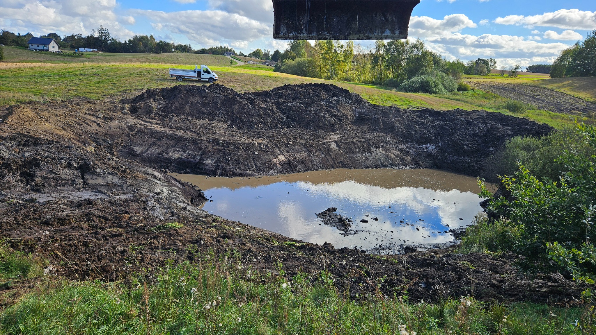 Wykopany staw z wodą i odbiciem nieba, na brzegu ziemia i roślinność, w tle biała ciężarówka i budynek. Nad wykopem fragment łyżki koparki.