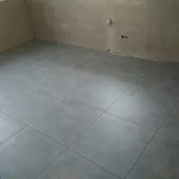 Świeżo ułożona, szara, matowa podłoga z płytek ceramicznych w pustym, surowym pomieszczeniu z widocznymi śladami po remoncie na ścianach.
