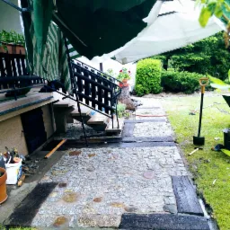 Fragment układanej ścieżki z kostki granitowej z drewnianymi obrzeżami, widoczne narzędzia, schody z czarną barierką i prowizoryczne zadaszenie.