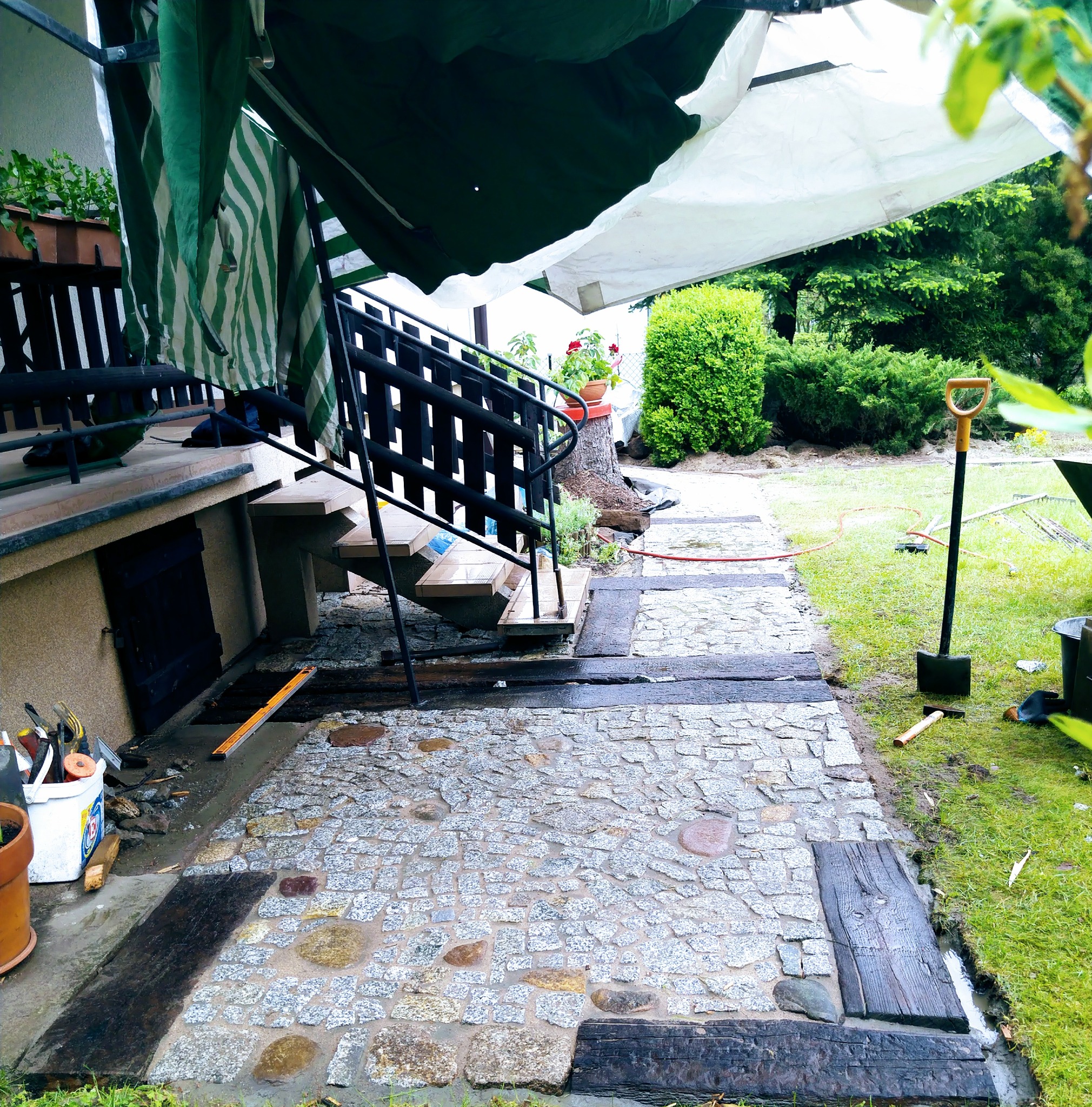 Fragment układanej ścieżki z kostki granitowej z drewnianymi obrzeżami, widoczne narzędzia, schody z czarną barierką i prowizoryczne zadaszenie.