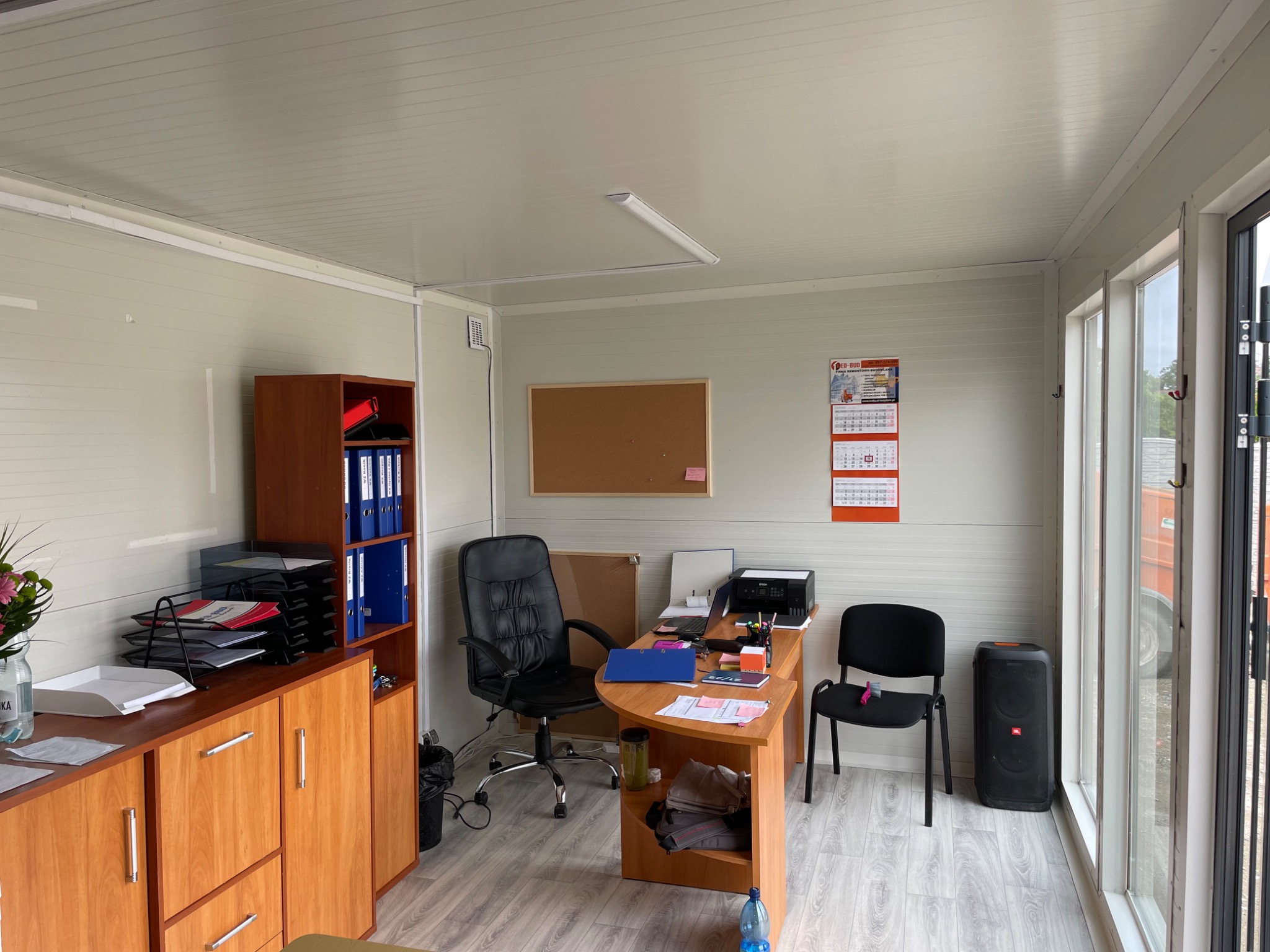 Wnętrze biura kontenerowego z drewnianą podłogą, wyposażone w biurko, krzesła, regał z segregatorami i sprzęt biurowy, oświetlone świetlówką.