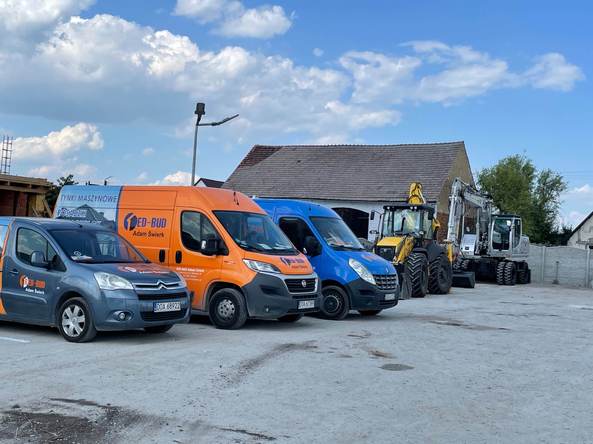 Flota pojazdów budowlanych: szara osobówka, pomarańczowy i niebieski van z logo firmy oraz koparki na betonowym placu pod błękitnym, pochmurnym niebem.