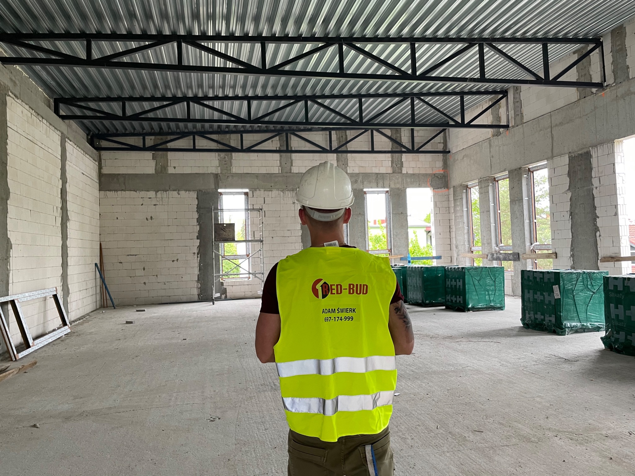 Wnętrze budynku w trakcie budowy, widoczne ściany z białych bloczków, stalowa konstrukcja dachu, betonowa podłoga i pracownik w kasku i kamizelce odblaskowej z logo firmy 'RED-BUD', stojący tyłem...