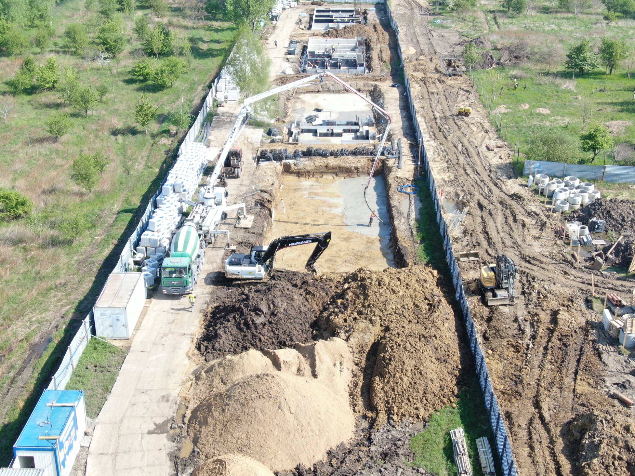 Widok z góry na plac budowy z wykopami, fundamentami w szalunkach, betoniarką, pompą do betonu i koparką HIROMEK pracującą przy hałdach ziemi.