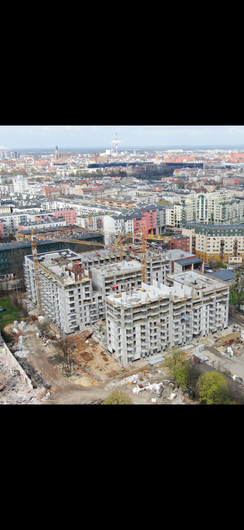 Wysoki kąt widokowy na plac budowy wielopiętrowego budynku mieszkalnego w trakcie realizacji, z widocznymi dźwigami i panoramą miasta w tle.
