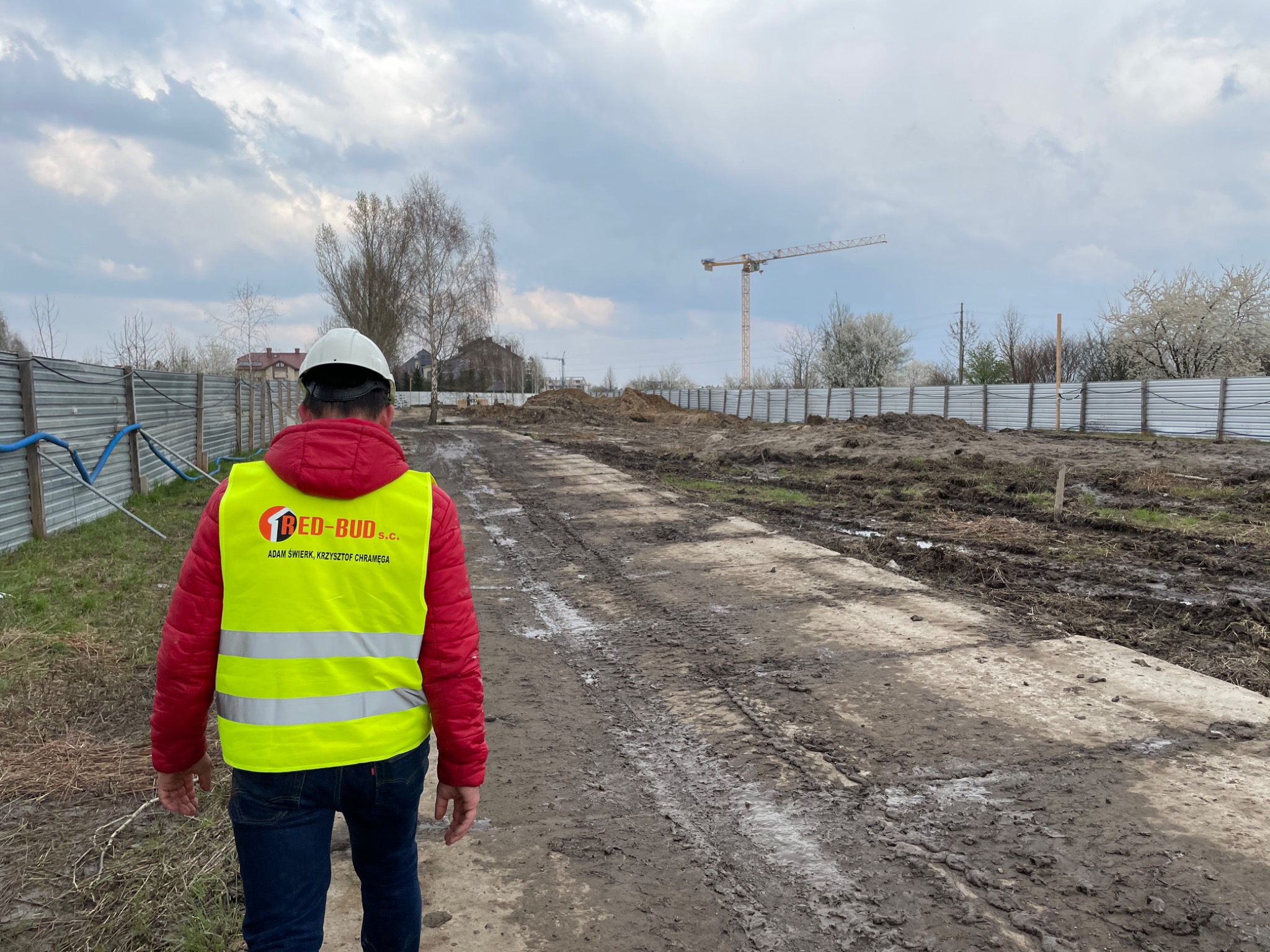 Pracownik w kasku i kamizelce odblaskowej z logo firmy budowlanej na terenie budowy, widok z tyłu, w tle żuraw budowlany i ogrodzenie z blachy falistej.