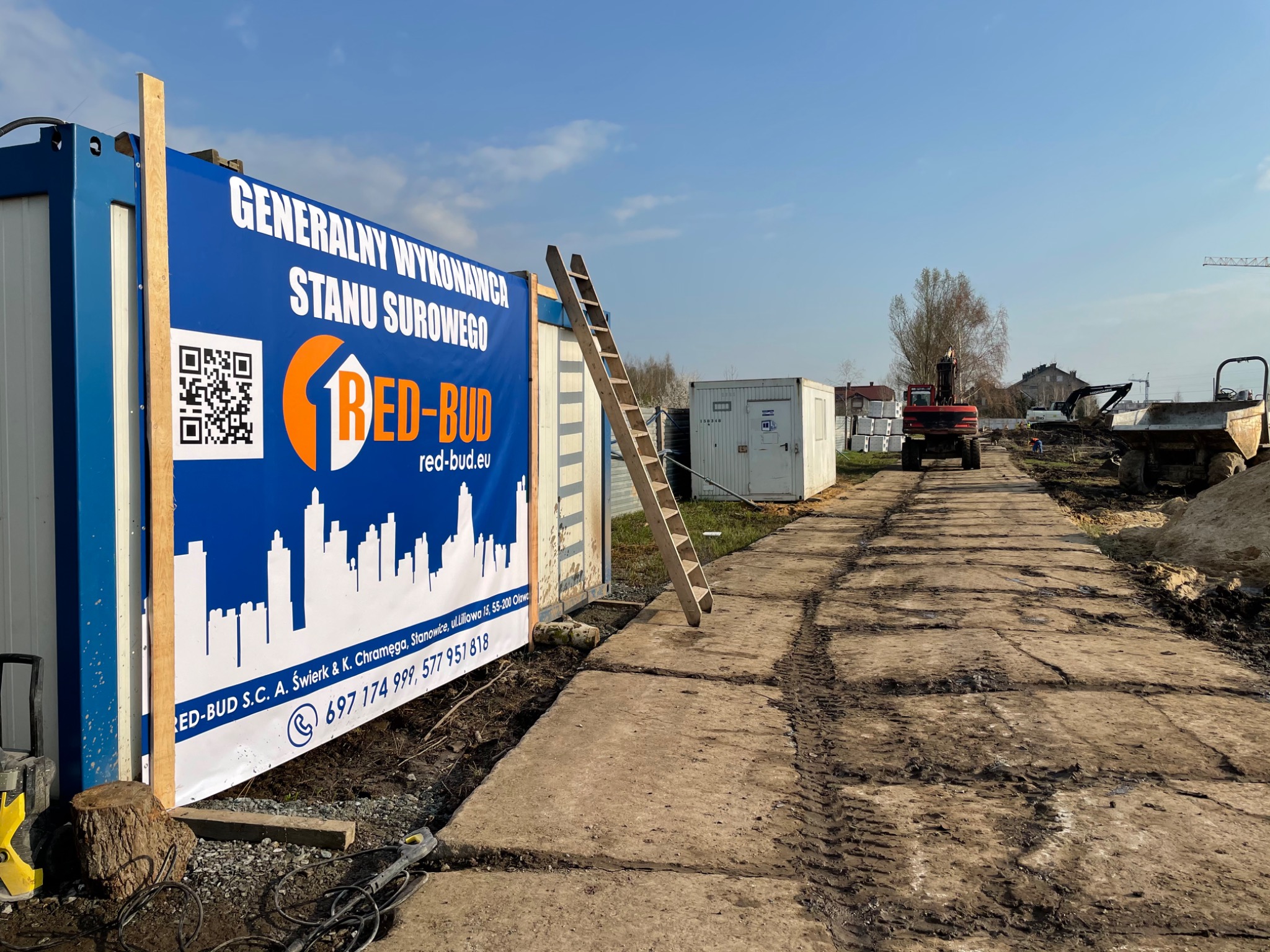 Plakat informacyjny firmy budowlanej RED-BUD na kontenerze budowlanym, obok drabina, w tle teren budowy z maszynami i kontenerami.