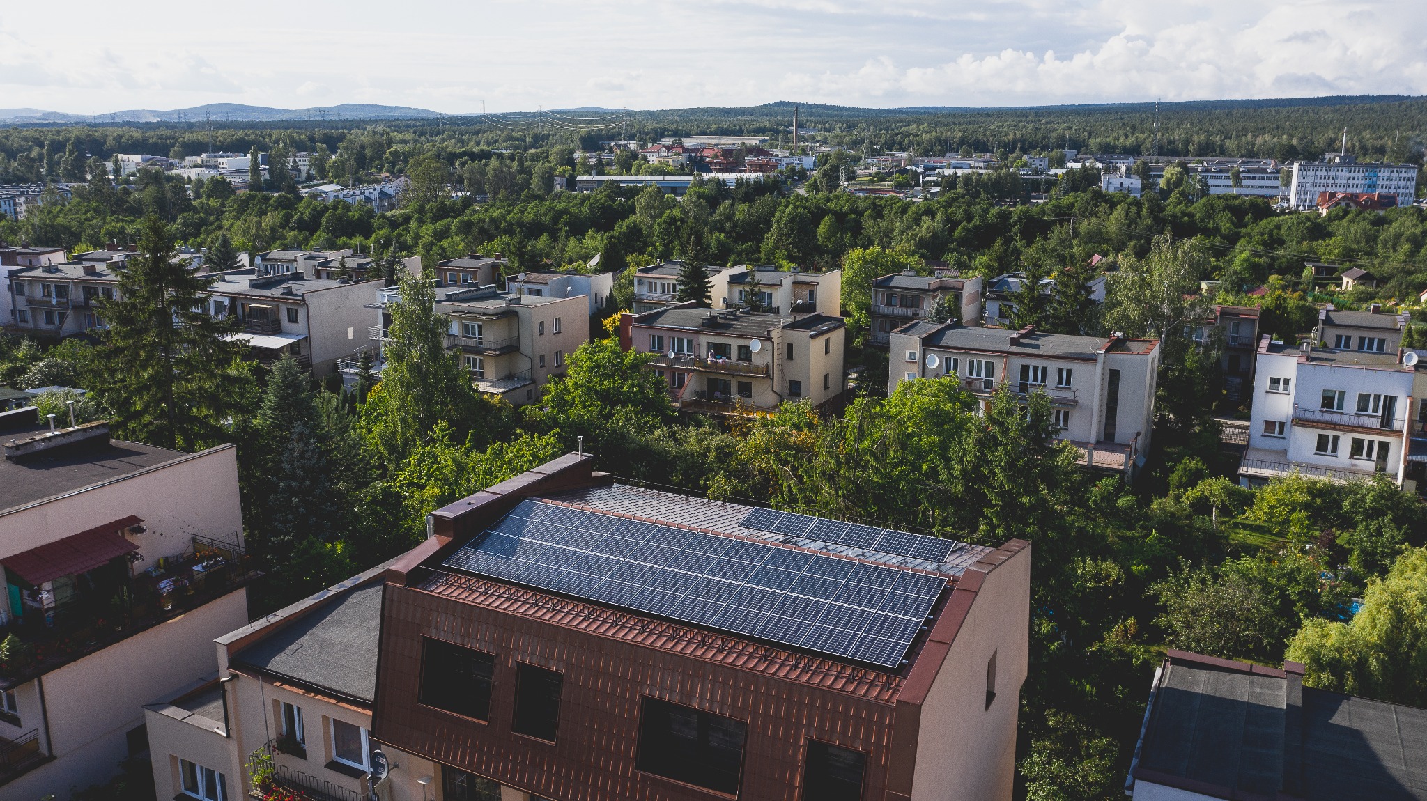 Panel słoneczny na dachu budynku mieszkalnego z widokiem na miasto w tle, zieleń drzew i błękitne niebo z chmurami.