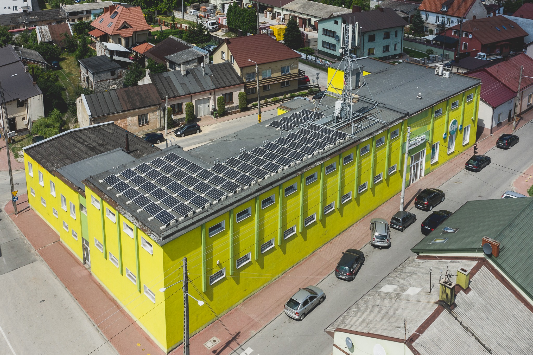 Widok z góry na budynek z żółtą fasadą i panelami fotowoltaicznymi na dachu, w tle zabudowa miejska i wieża telekomunikacyjna.