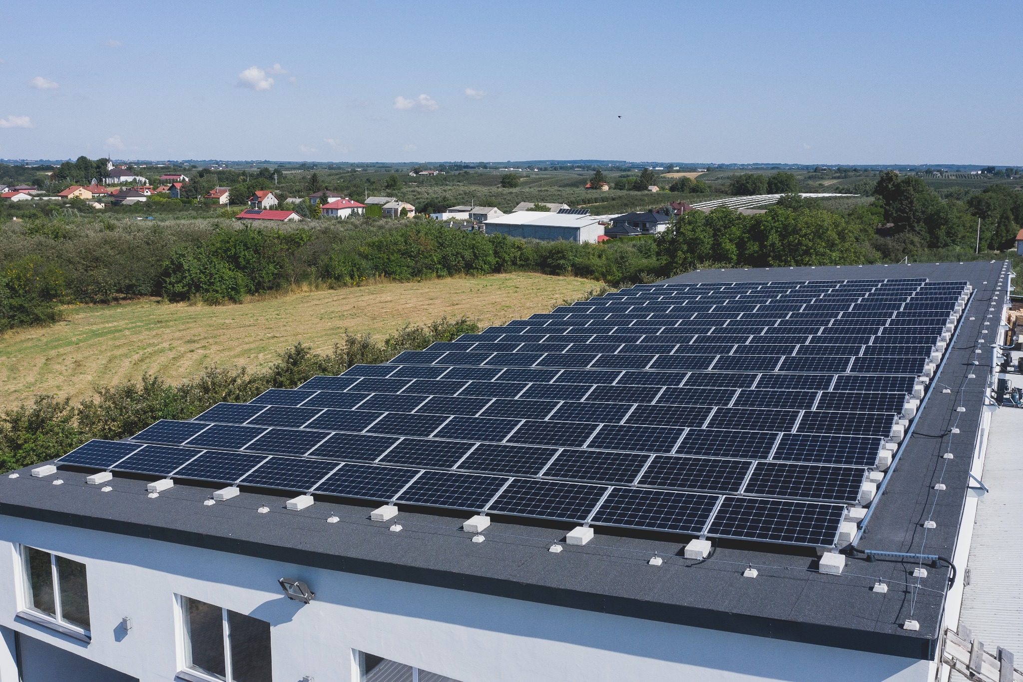 Widok z góry na dużą instalację paneli słonecznych na płaskim dachu budynku, z widocznymi elementami mocującymi i przewodami, na tle wiejskiej zabudowy i błękitnego nieba.