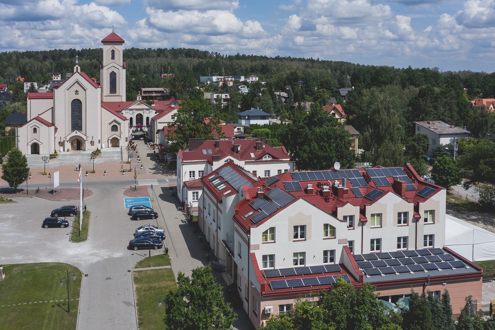 Widok z góry na budynki z czerwonymi dachami, na których zamontowane są panele słoneczne, w tle kościół i drzewa.