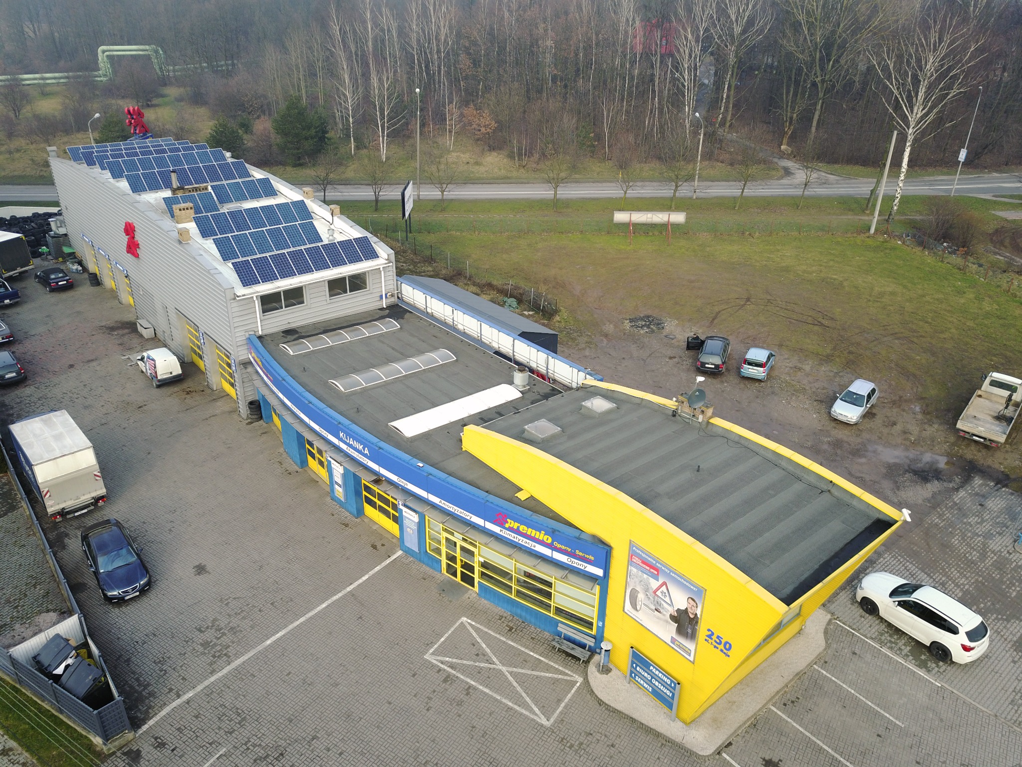 Widok z góry na budynek serwisu samochodowego Premio z panelami słonecznymi na dachu, zaparkowane samochody i otaczający teren z drzewami.