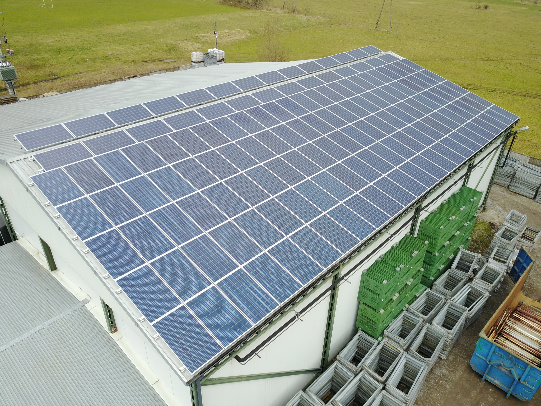 Widok z góry na halę z panelami słonecznymi na dachu, obok składowane puste metalowe ramy i zielone skrzynki.