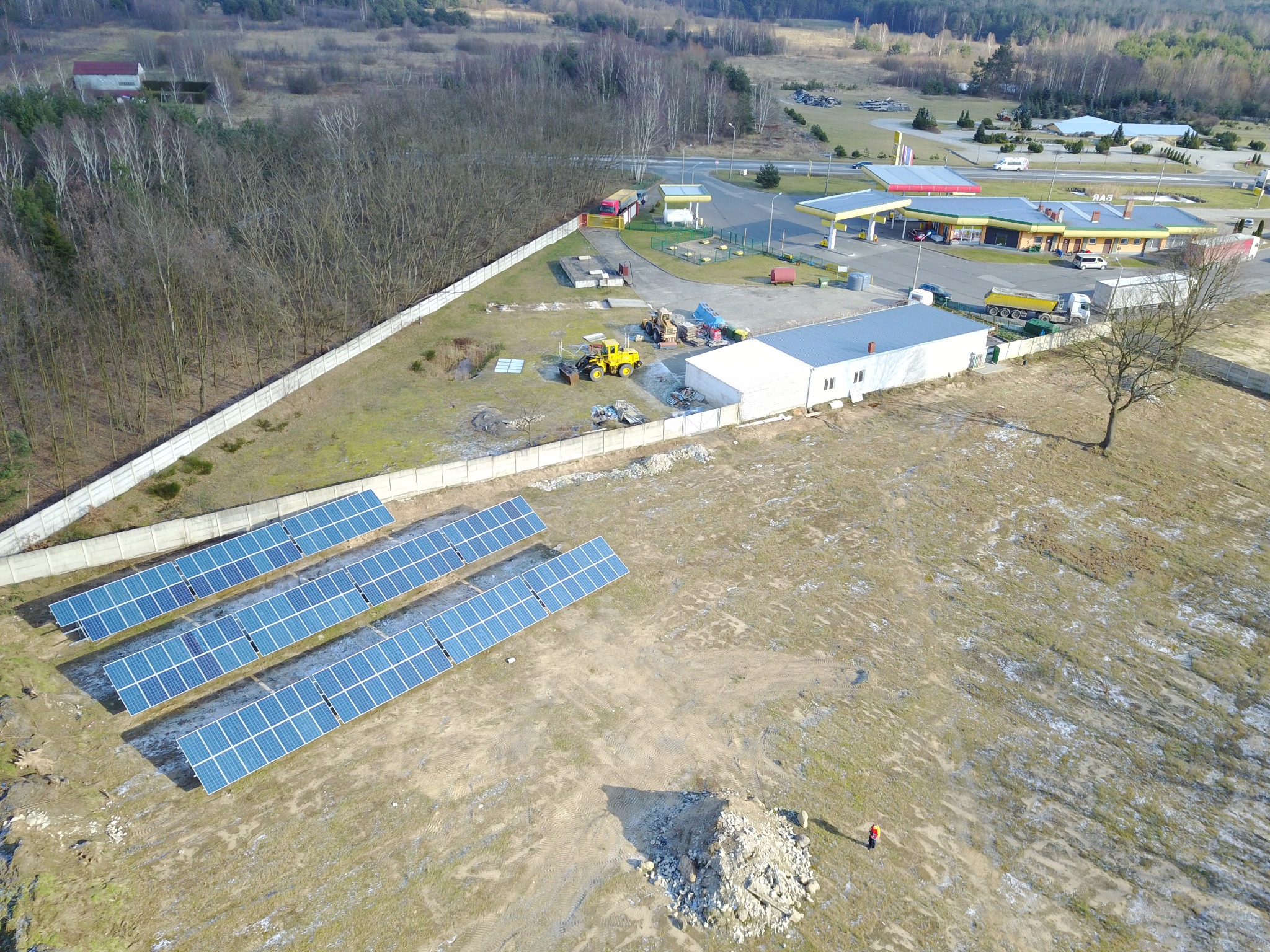 Widok z drona na instalację paneli słonecznych na polu, z widoczną stacją benzynową w tle i pracującymi maszynami budowlanymi w pobliżu.