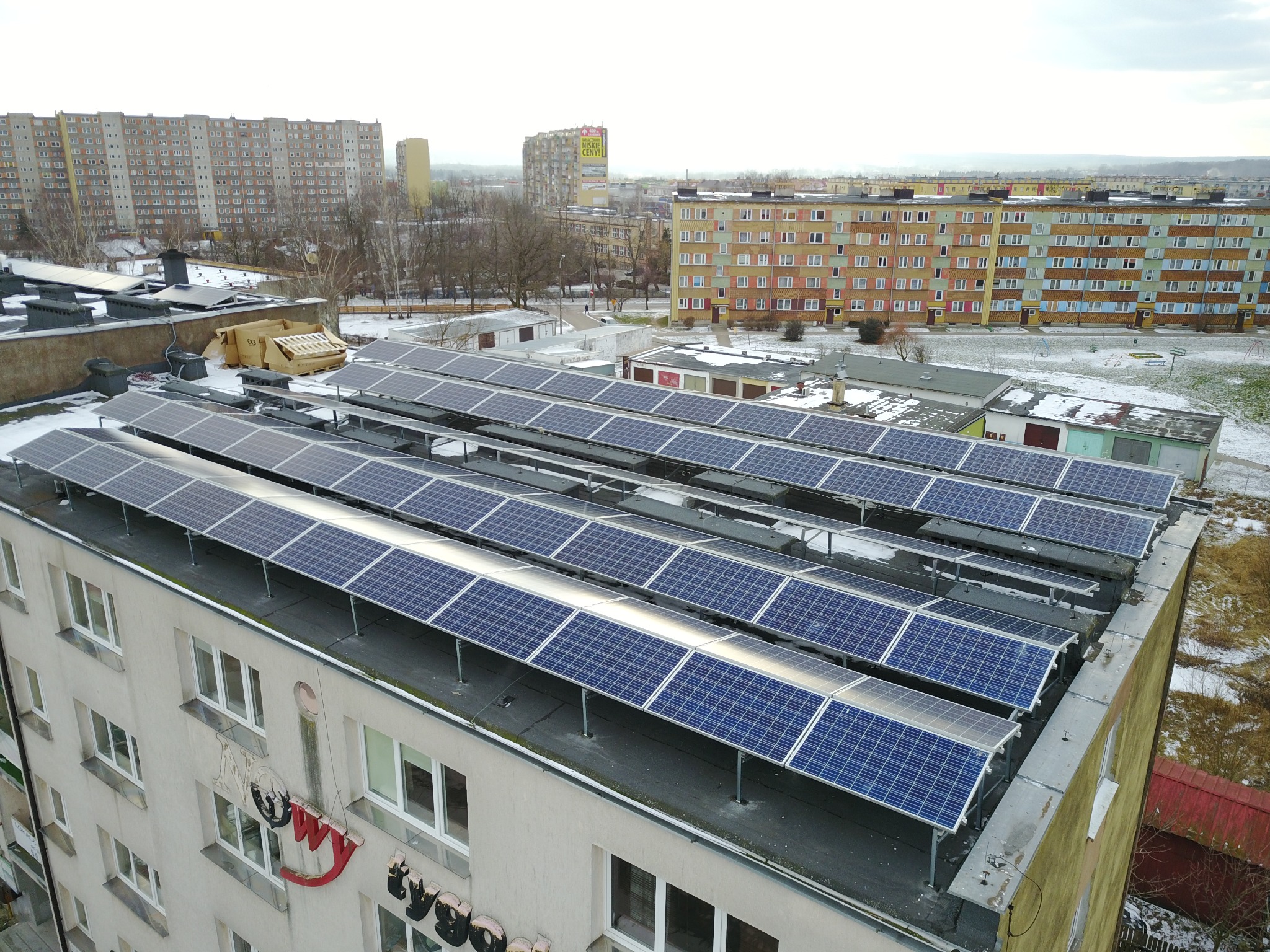 Rząd paneli słonecznych na dachu budynku mieszkalnego, widok z góry, w tle zabudowa miejska i lekko pochmurne niebo.