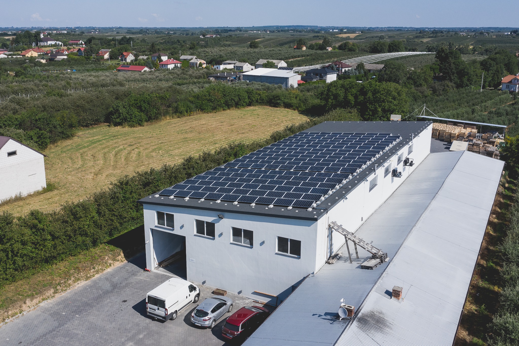Widok z góry na budynek przemysłowy z panelami fotowoltaicznymi na dachu, zaparkowane samochody i otaczający krajobraz wiejski.
