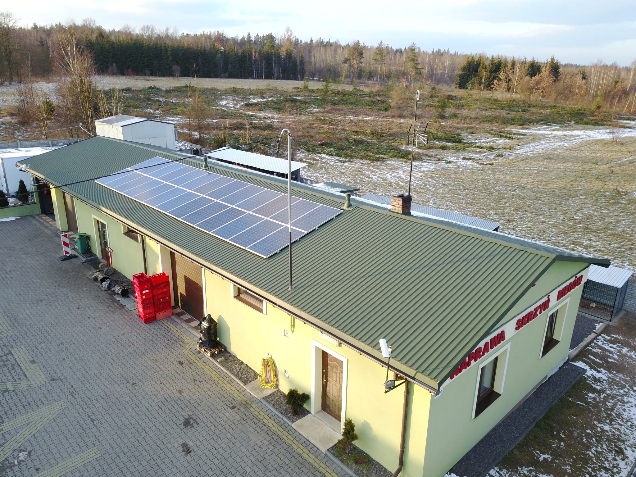 Widok z góry na budynek z panelami słonecznymi na dachu, z widocznymi elementami otoczenia: drzewa, częściowo ośnieżone pole, kostka brukowa przed budynkiem oraz czerwone skrzynki.