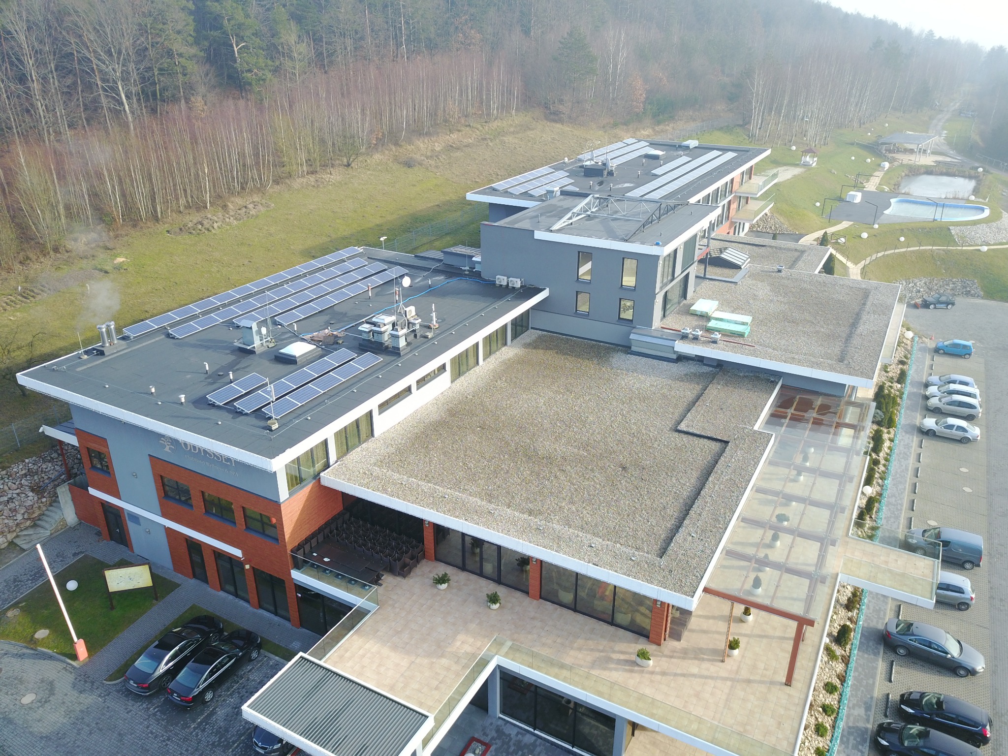 Widok z lotu ptaka na budynek hotelu z panelami słonecznymi na dachu, w tle las i basen.