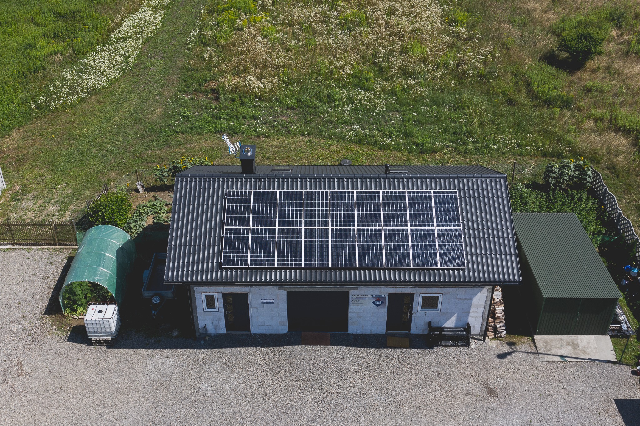Widok z góry na budynek z panelami słonecznymi na dachu, obok szklarnia tunelowa i zagospodarowany teren zielony.