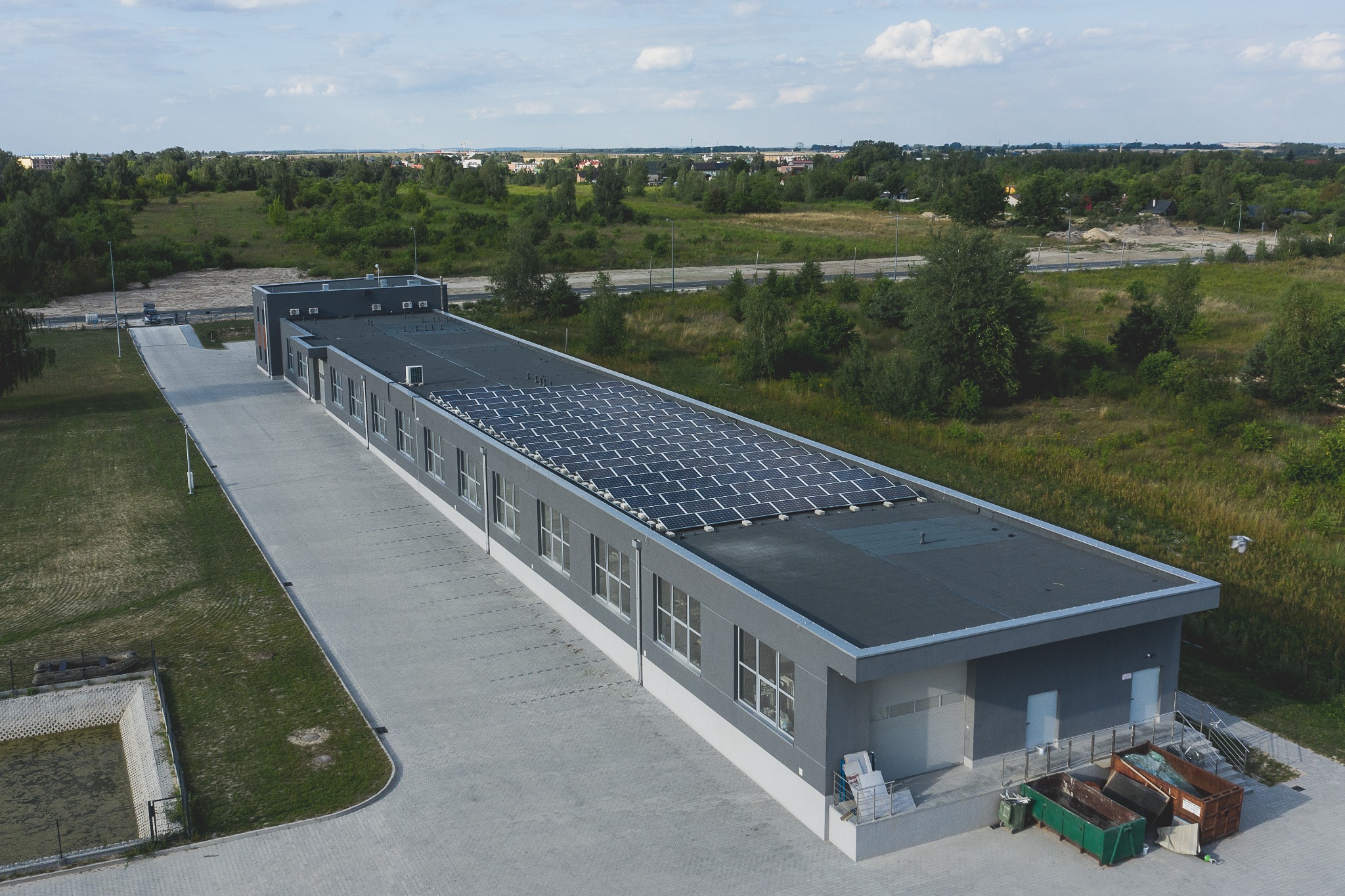 Widok z góry na budynek przemysłowy z panelami słonecznymi na dachu, otoczony zielenią i betonowym placem