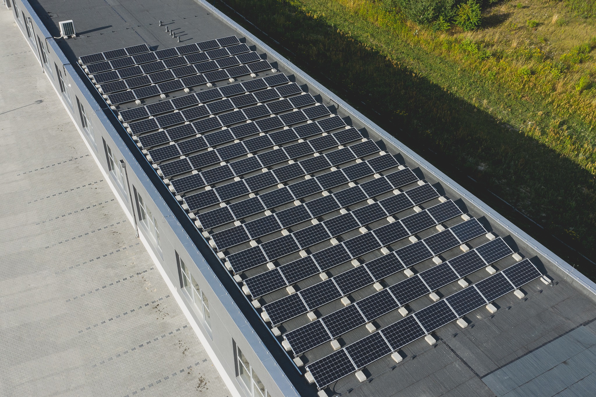 Widok z góry na dużą instalację fotowoltaiczną na płaskim dachu budynku przemysłowego, z widocznymi rzędami paneli słonecznych i zielonym tłem roślinności w oddali.