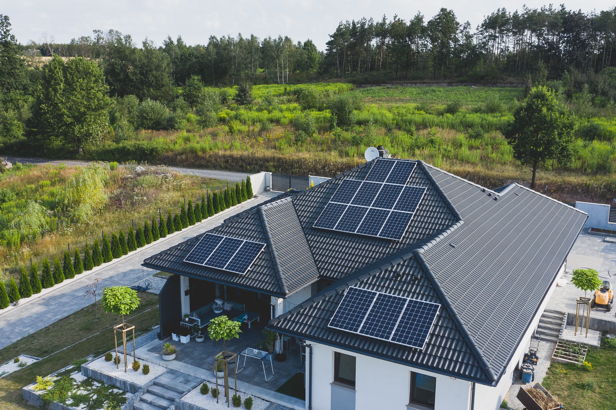 Widok z lotu ptaka na dom jednorodzinny z ciemną dachówką, na której zamontowano panele słoneczne; w tle zielone drzewa i krzewy.