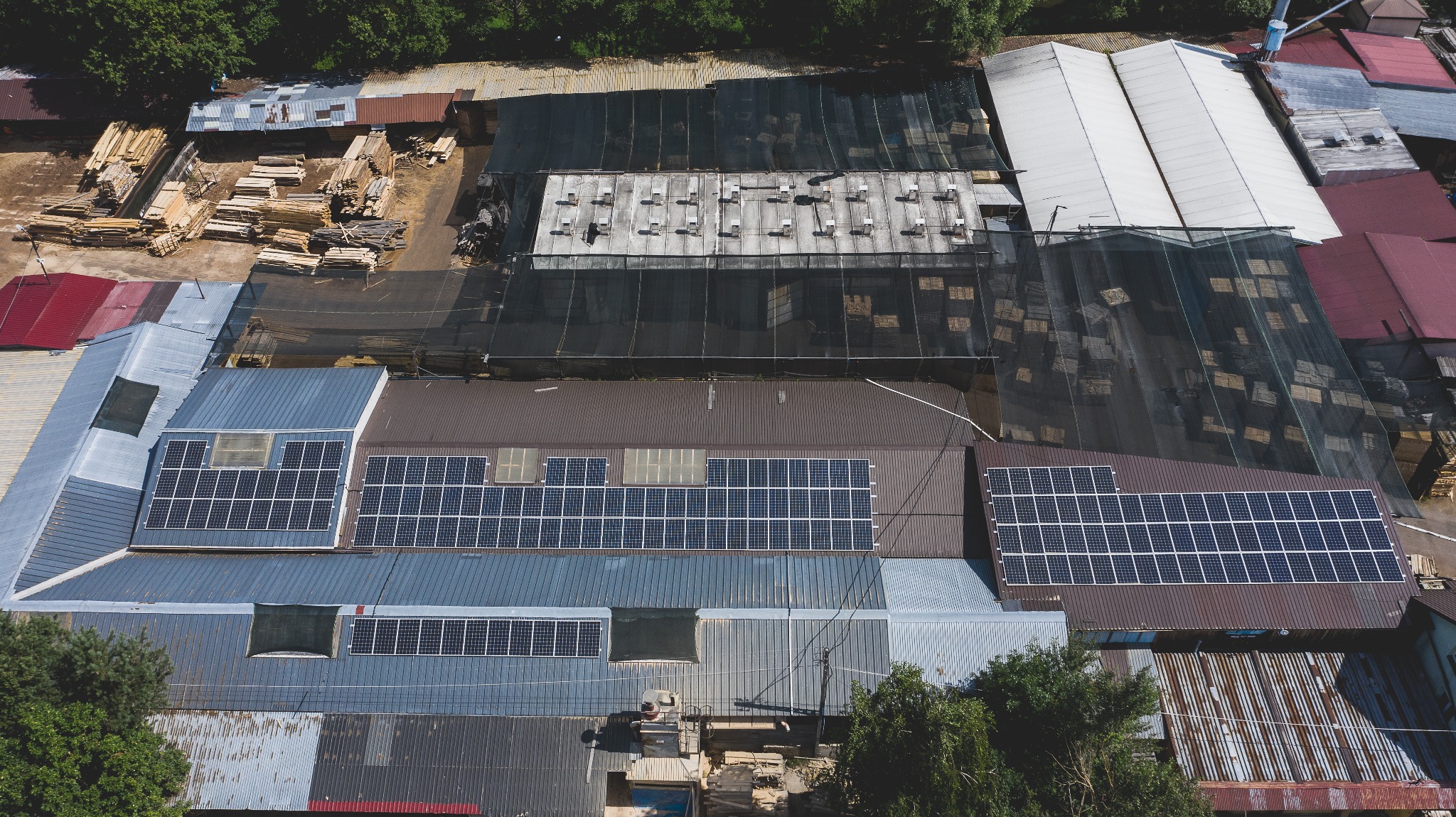 Widok z góry na dachy budynków z zainstalowanymi panelami słonecznymi, obok składowane drewno i zadaszone magazyny.