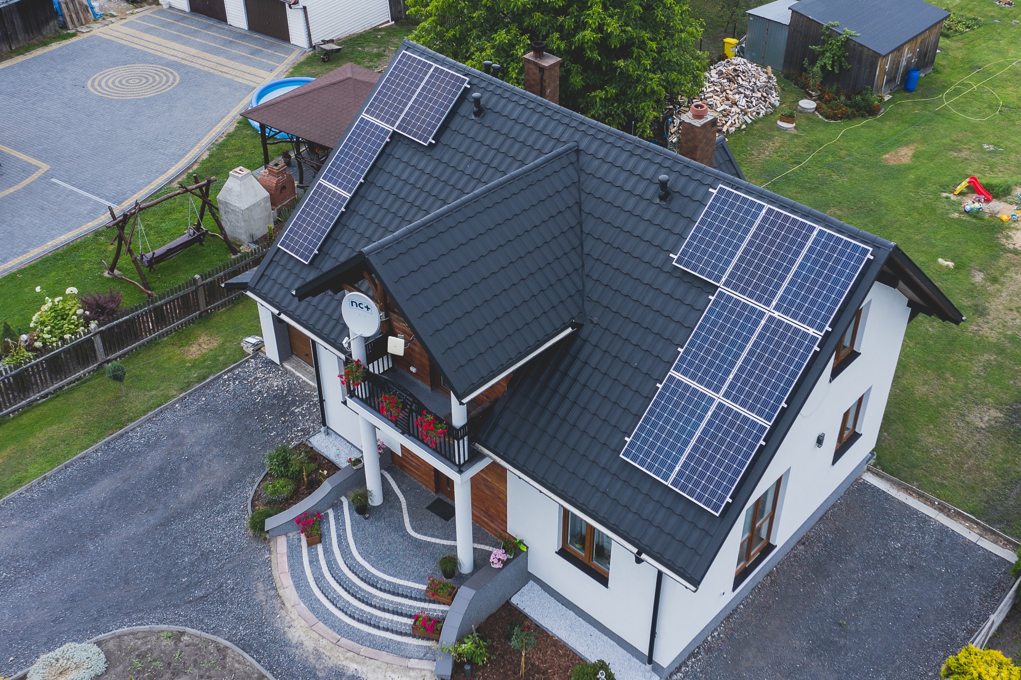Widok z góry na dom jednorodzinny z zainstalowanymi panelami słonecznymi na czarnym dachu, widoczna antena satelitarna na elewacji, ozdobny ogród z kamiennymi schodami i kwiatami.