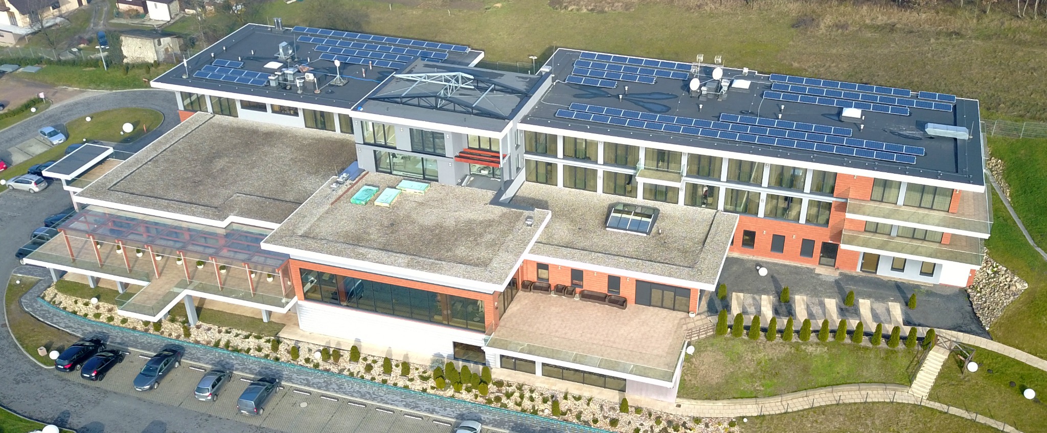 Widok z lotu ptaka na duży budynek z panelami słonecznymi na dachu, otoczony zielenią i parkingiem z kilkoma samochodami.