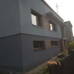 Dwukondygnacyjny dom z szarą elewacją, fragment ściany z oknami i dekoracyjną cegłą, widok z niskiej perspektywy, w tle sąsiedni budynek i fragment drewnianego płotu.